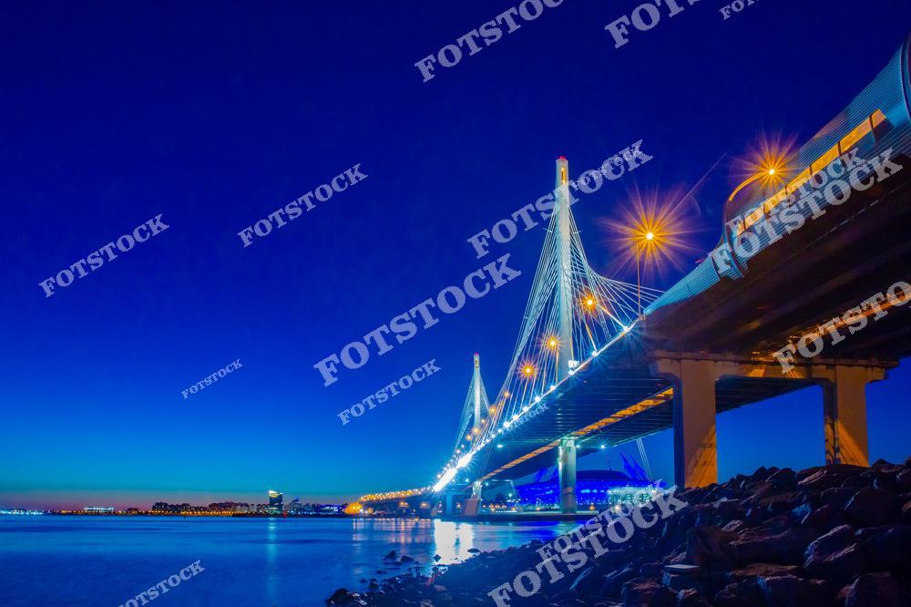 Фотография ночного Санкт-Петербург, Россия. На изображении показан вантовый Обуховский мост, освещённый огнями ночного города. Фотоснимок подчёркивает современную архитектуру Петербурга: мощные опоры, вантовые конструкции и отражения света на фоне тёмного неба. Фото передаёт атмосферу ночного Петербурга и демонстрирует инженерные достижения мостостроения. Иллюстрация может использоваться как изображение, картинка или слайд, посвящённые мостам Санкт-Петербурга, архитектуре и городам России.